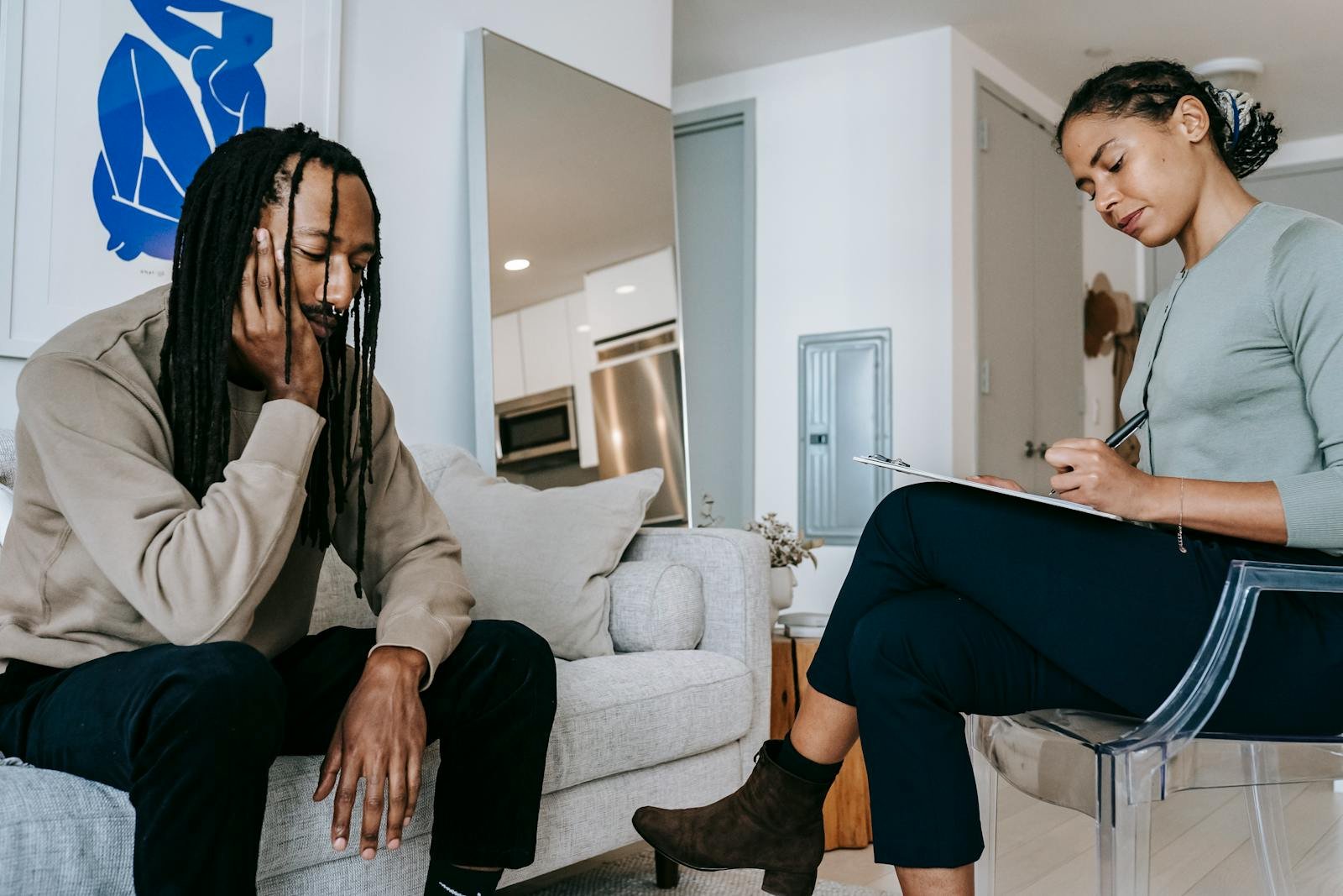Attentive African American female with clipboard taking notes near sad male partner touching face on couch at home